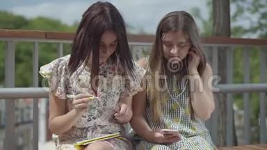 两个漂亮的女学生坐在户外。 一个女孩带着耳机听音乐，第二个女孩